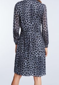 Kobieta w sukience w panterkę, do kolan, z długim rękawem, widziana od tyłu na tle jednolitym.