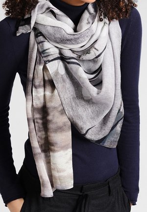 Femme portant une grande écharpe gris clair à motif, drapée sur une chemise à manches longues bleu marine foncé et un pantalon noir.