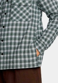 Camisa de franela a cuadros verdes y blancos con botones negros, un bolsillo en el pecho y un corte relajado. La tela tiene una apariencia texturizada.