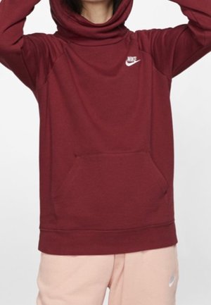 Person trägt einen dunkelroten Nike-Hoodie mit einer vorderen Tasche und blassrosa Nike-Sweathosen, Arme über dem Kopf erhoben.