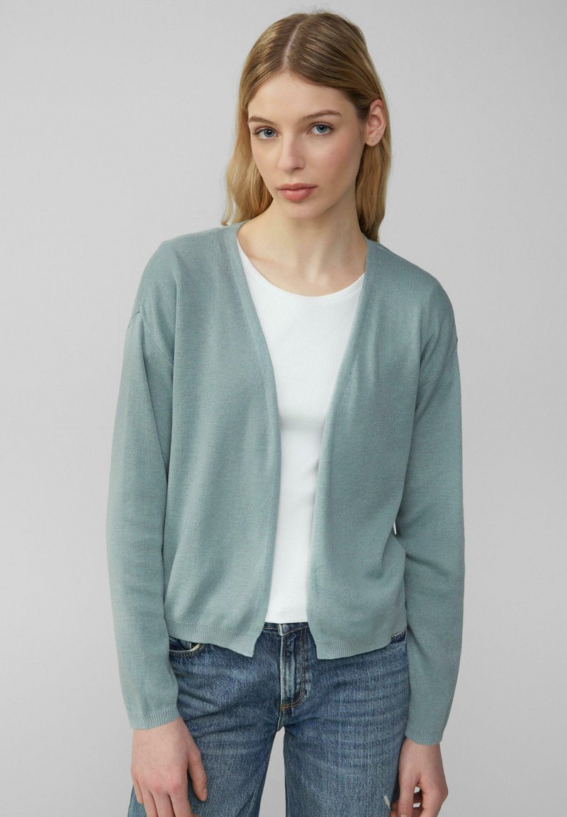 Hellgrüner Cardigan mit entspannter Passform, offenem Vorderteil und langen Ärmeln, getragen über einem weißen Oberteil. Glatte Textur und minimale Details.