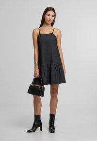 Melna bezsvārku kleita ar pakāpenisku apakšmalu, izgatavota no teksturēta auduma. Modelis tur mazs melns rokassomu; valkā melnas kurpes līdz potītei.