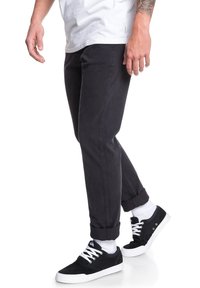 Pantalon en coton noir avec une coupe fuselée, retroussés aux chevilles, associés à des baskets noires et des chaussettes blanches. Le tissu est lisse avec une légère texture.