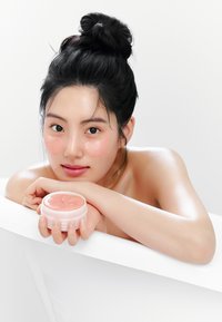 Crème de soin rose dans un récipient rond et transparent tenu dans une main. Texture lisse visible. Fond épuré et clair mettant en valeur le produit.