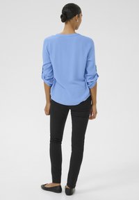 Chemise à manches longues bleu clair texturée avec les manches retroussées, associée à un jean skinny noir et des ballerines noires. Vue de dos.