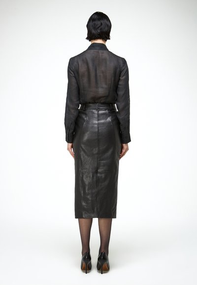 Žena stojaca chrbtom ku kamere, oblečená v priesvitnej čiernej blúzke, čiernej ceruzkovej sukni z kože s vysokým pásom, priesvitných pančuchách a čiernych lodičkách na vysokom opätku.