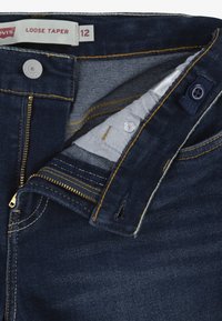 Jean Levi's à coupe large et fuselée, bleu foncé, taille 12, avec fermeture éclair dorée partiellement ouverte, bouton apparent et détails du tissu intérieur visibles.