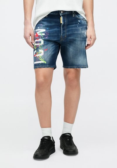 Shorts en jean bleu foncé avec un ourlet effiloché, arborant un motif éclaboussé de peinture vibrant et un texte blanc sur la jambe gauche. Baskets noires portées.