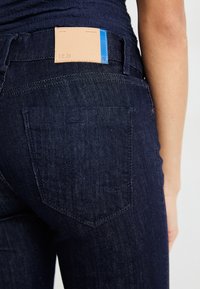 Tmavě modré džíny z denimu mají hladkou texturu, výrazné zadní kapsy a hnědou koženou nášivku s modrým akcentem na pase.