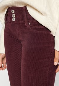 Burgundské manžestrové kalhoty se zapínáním na tři knoflíky, texturovaná látka a decentní detaily kapes. V kombinaci s krémovým svetr.