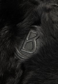 Schwarzer Pelzstoff mit einem strukturierten Lederpatch, der mit einem stilisierten "B" geprägt ist. Der Pelz hat ein weiches, dichtes Erscheinungsbild.