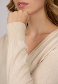 Pull en tricot beige avec un col en V. Dispose de poignets côtelés et d'une texture douce, mettant en avant un design léger et confortable.