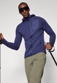 Veste zippée bleu marine avec une finition texture, dotée d'une capuche, de deux poches zippées et d'un panneau réfléchissant sur la poitrine. Portée avec un pantalon beige.
