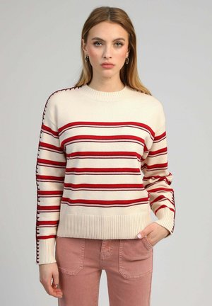 Femme portant un pull crème avec des rayures horizontales rouges et un pantalon rose, debout devant un fond gris clair uni.