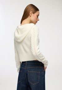 Biała krótkotrwała bluza z kapturem, wykonana z teksturowanego dzianiny, z kapturem i rękawami z warkoczy, które mają wycięcia. W połączeniu z ciemnoniebieskimi dżinsami z wysokim stanem.