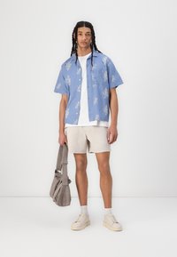 Chemise courte à manches bleues claires avec des motifs floraux blancs, t-shirt blanc en dessous, shorts beiges, baskets beiges et un sac à bandoulière gris.