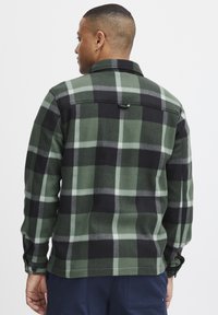 Camisa de franela a cuadros verde y negra con cuello, puños abotonados y un lazo colgante en la parte de atrás; textura suave en un corte ajustado.