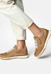 Baskets en suedine beige clair avec des lacets blancs, détail de fermeture éclair sur le côté et semelle en caoutchouc texturée. Portées avec un pantalon clair retroussé.
