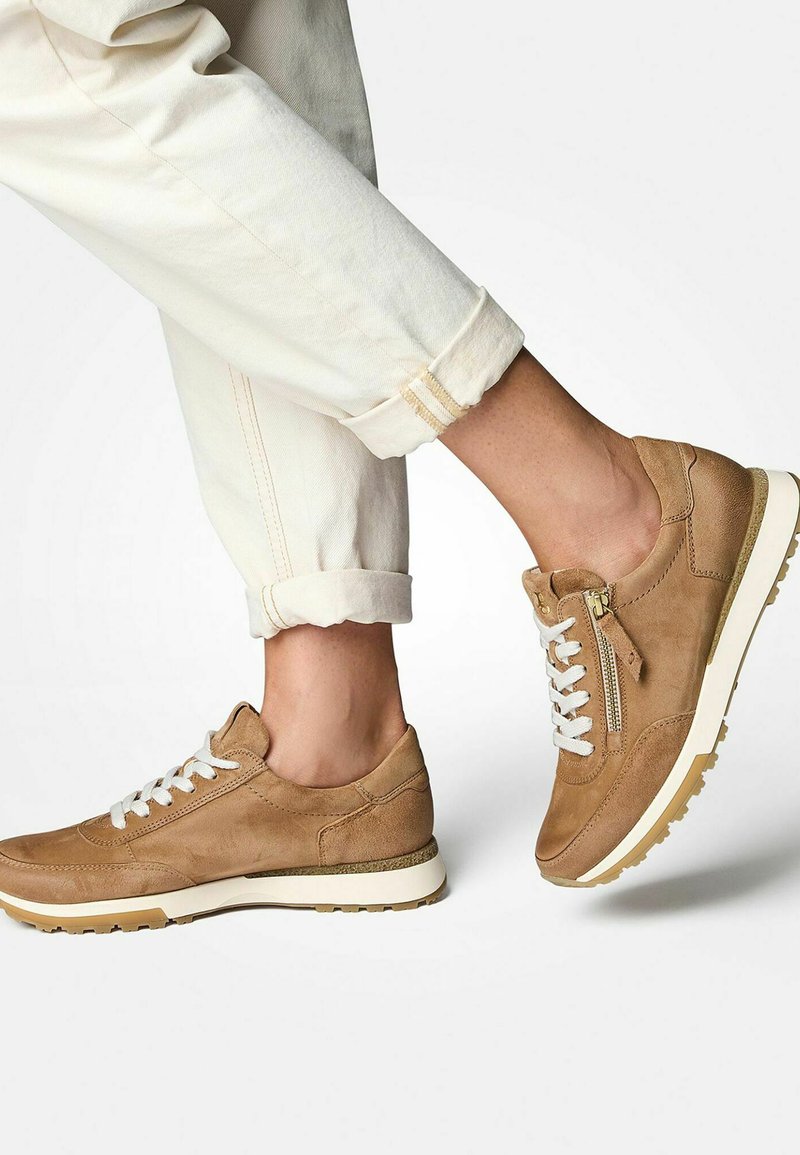 Baskets en suedine beige clair avec des lacets blancs, détail de fermeture éclair sur le côté et semelle en caoutchouc texturée. Portées avec un pantalon clair retroussé.