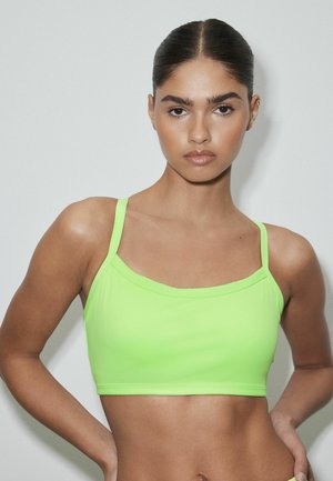 Junge Frau mit sleek zurückgebundenem Haar, die ein neon-grünes, bauchfreies Top trägt, steht vor einem schlichten hellen Hintergrund.