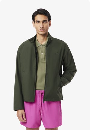 Olivegrünes, leichtes Jackett mit hohem Kragen und Reißverschluss, getragen über einem hellgrünen Poloshirt, kombiniert mit knallpinken Shorts.