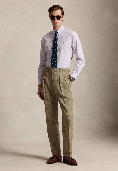 Muž so slnečnými okuliarmi, pruhovanou košeľou na gombíky, vzorovanou kravatou, vysokým pásom károvaných nohavíc, hnedými mokasínmi a náramkovými hodinami na jednoduchom pozadí.