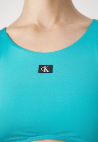 Türkiz színű, testhezálló ujjatlan felső, sima textúrával, fekete téglalap alakú címkével, amelyen a nyakvonal közelében fehér betűkkel az áll: "cK".