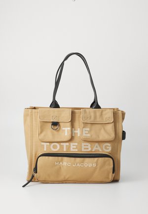 Beige canvas-kassi mustilla kahvoilla, joissa on kaksi etutaskua läpällä, vetoketjutasku ja teksti "THE TOTE BAG MARC JACOBS".
