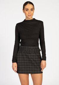 Vrouw in een zwarte langemouwen top en een hoog-gesneden minirok van donker plaid met een brede riem, staande tegen een witte achtergrond.