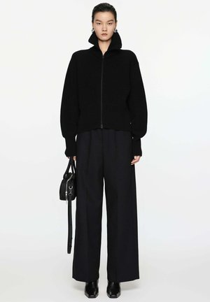 Schwarze Strickjacke mit Reißverschluss und hohem Kragen, kombiniert mit lockeren schwarzen Hosen und eleganten schwarzen Stiefeletten. Inklusive einer kleinen schwarzen Tasche.