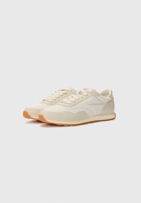 Sneakers feature a light beige textile upper with suede accents, rounded toe, and a gum rubber sole. Minimalist design and lace-up closure.