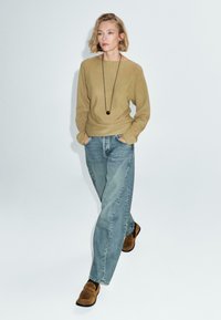 Frau in beigem Pullover, blauen Jeans und braunen Wildlederschuhen, die mit den Händen in den Taschen vor einem schlichten Hintergrund geht.
