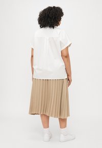 Blouse blanche à manches courtes avec un détail de découpe au dos, associée à une jupe midi plissée beige. Chaussures : baskets blanches avec des chaussettes montantes.