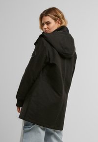 Manteau noir à capuche, avec une coupe décontractée, doté de poignets côtelés, de poches latérales et d'une texture lisse. L'arrière présente un détail de patch logo.