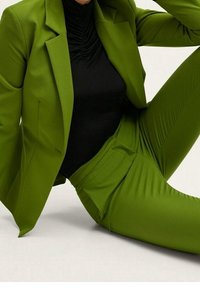 Personne portant un costume vert vif avec un pantalon et un blazer sur mesure, associé à un haut noir froncé, assise devant un fond neutre.