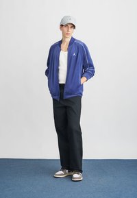 Blå velour zip-up jakke med hvite detaljer, hvit tank topp, svarte bukser, grå cap og brune sneakers med fargerike lisser.