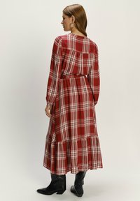 Dlouhé červené kostkované šaty s dlouhými rukávy, nařasenými rameny a vícevrstvou sukní, doplněné černými kotníkovými botami.