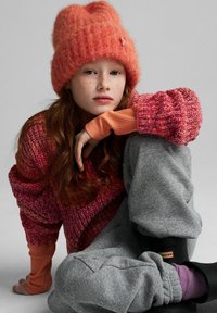 Pomarańczowa dziergana czapka, żywy sweter o fakturze w odcieniach czerwieni i różu, szare spodnie dresowe oraz czarne buty z detalem logo.