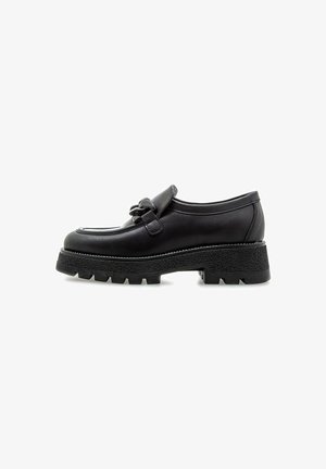 Zapato de cuero negro con punta redondeada, una correa decorativa y una suela de goma gruesa y texturizada. Diseño minimalista con sutiles detalles de costura.