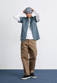 Ριγέ πουκάμισο σε ναυτικό και λευκό χρώμα, φορεμένο πάνω από λευκό μπλουζάκι με μακριά μανίκια. Καφέ cargo παντελόνι και μαύρα αθλητικά παπούτσια με λευκή σόλα. Πλεκτό σκουφί.