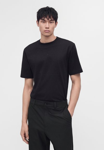 Jeune homme aux cheveux courts et noirs portant un t-shirt noir uni et un pantalon noir, debout avec une main dans la poche devant un fond uni.