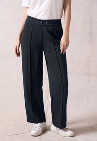 Pantaloni neri a gamba larga con una texture liscia, frontale plissettato e vita elastica aderente. Abbinati a sneakers bianche.