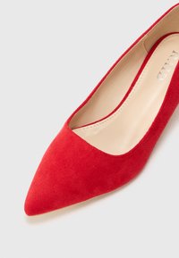 Pumps tinggi merah suede dengan ujung runcing dan interior halus berwarna krem. Desainnya menampilkan vamp yang rendah untuk siluet yang ramping.