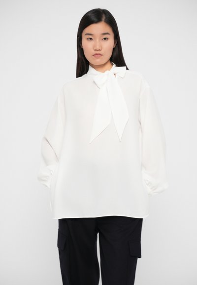 White blouse with a bow tie collar, long puffed sleeves, and a relaxed fit. Smooth fabric texture, subtle sheen. Paired with dark pants.