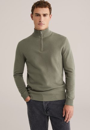 Pulover verde tricotat cu guler cu jumătate de fermoar, țesătură texturată, manșete și tiv din material rib bată. Purta cu blugi negri.