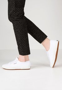 Hvite kanvas sneakers med flate såler, runde tær og teksturerte gummisåler, sammen med svarte ankelsbukser med prikker, føttene i bevegelse.