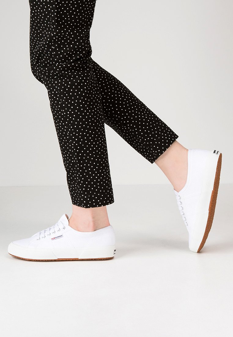 Hvite kanvas sneakers med flate såler, runde tær og teksturerte gummisåler, sammen med svarte ankelsbukser med prikker, føttene i bevegelse.