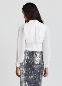 Blusa bianca con scollo arricciato e maniche lunghe leggermente gonfie, abbinata a una gonna di paillettes argentate che riflettono luce e texture.