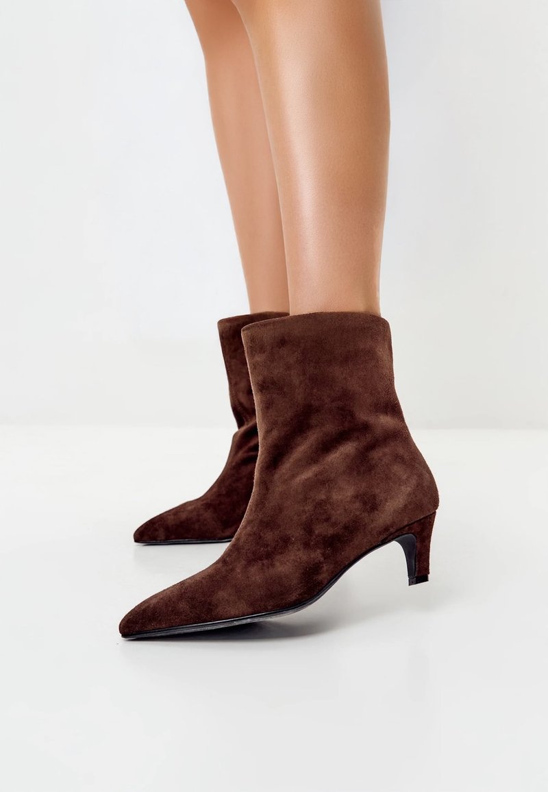 Barna színű, velúr bokacsizma hegyes orral és alacsony, keskeny sarokkal. Simább textúrájú, testhez simuló kialakítás, hajtókás mandzsettával.