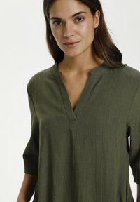 Olijfgroene blouse met een V-hals en driekwart mouwen. Gemaakt van textiel met structuur, met een ontspannen pasvorm en subtiele stikseldetails.